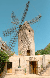 Un antiguo molino de piedra rústico con grandes aspas se alza contra el cielo despejado, mostrando el encanto de Mallorca. Explora su interior y participa en el "Taller de cocina mediterránea en Mallorca" para disfrutar de una vibrante experiencia culinaria mediterránea. - ForTwo - Planes para parejas