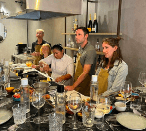 En Taller Mediterráneo de Sabores en Palma, la gente prepara la comida con alegría en una cocina animada, mezclando sabores vibrantes con una variedad de ingredientes y utensilios esparcidos por el mostrador. - ForTwo - Planes para parejas