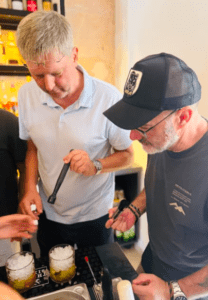 Dos personas mezclan bebidas con destreza en un bar, una de ellas con una herramienta en el bullicioso Taller de coctelería en Cala d’Or, haciendo eco de las vibrantes noches de Cala d’Or. - ForTwo - Planes para parejas