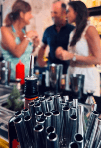 Una escena animada se desarrolla mientras la gente charla en un bar de Cala d’Or, con pajitas de cóctel en foco, evocando la esencia del Taller de coctelería en Cala d’Or. - ForTwo - Planes para parejas