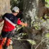 Equipado con material de seguridad, un escalador se enfrenta al terreno rocoso de La Cantera, agarrado a una cuerda en un estrecho paso del acantilado durante una emocionante experiencia en la Vía Ferrata de Iniciación en La Cantera. - ForTwo - Planes para parejas