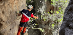 Equipado con material de seguridad, un escalador se enfrenta al terreno rocoso de La Cantera, agarrado a una cuerda en un estrecho paso del acantilado durante una emocionante experiencia en la Vía Ferrata de Iniciación en La Cantera. - ForTwo - Planes para parejas