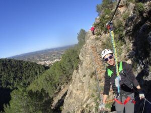 Sonriendo a la cámara, una persona recorre una montaña boscosa cerca de la Vía Ferrata en Fuente de Godalla, Valencia, equipada con equipo de seguridad para una aventura emocionante pero segura. - ForTwo - Planes para parejas