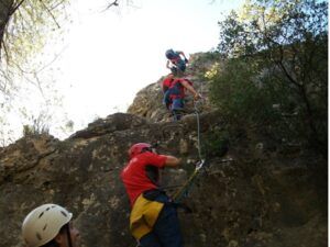 Con cascos, tres escaladores ascienden por un acantilado rocoso con cuerdas, árboles a su alrededor y un cielo despejado encima, enfrentándose a los desafíos de la Vía Ferrata en Fuente de Godalla - Valencia. - ForTwo - Planes para parejas