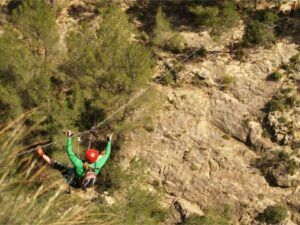 El aventurero, ataviado con un casco rojo y una chaqueta verde, asciende por un terreno accidentado que recuerda a la emocionante Vía Ferrata en Fuente de Godalla - Valencia, elevándose sobre un paisaje sembrado de árboles. - ForTwo - Planes para parejas