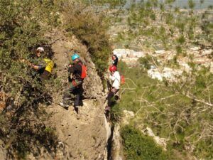 Cuatro escaladores, equipados con cascos y arneses, se enfrentan a los escarpados acantilados de la Vía Ferrata en Fuente de Godalla, Valencia. Rodeados de árboles, disfrutan de impresionantes vistas del pueblo a lo lejos. - ForTwo - Planes para parejas
