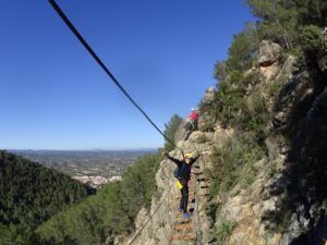 Una persona navega por la Vía Ferrata en Fuente de Godalla - Valencia con un arnés de seguridad bajo un cielo azul claro, encarnando la aventura. - ForTwo - Planes para parejas
