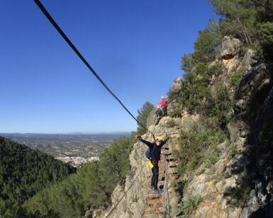 Una persona navega por la Vía Ferrata en Fuente de Godalla - Valencia con un arnés de seguridad bajo un cielo azul claro, encarnando la aventura. - ForTwo - Planes para parejas