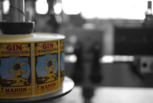 Las etiquetas de ginebra con diseño de molino de viento se despliegan en un entorno industrial difuminado, que recuerda a la Visita y Cata en Destilería de Mahón. - ForTwo - Planes para parejas