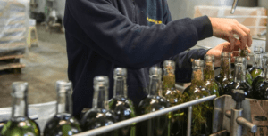 En una fábrica con mucho movimiento, un empleado introduce hierbas en botellas de vino con esmero, evocando la artesanía de la Destilería de Mahón. Los visitantes pueden disfrutar de la "Visita y Cata en la Destilería de Mahón", que desvela el arte detrás de cada sabor único. - ForTwo - Planes para parejas