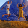 Dos personas saltan alegremente frente a un globo aerostático azul desinflado en un campo de hierba al atardecer, encarnando la esencia mágica de la experiencia Vuelo en globo en Valencia. - ForTwo - Planes para parejas