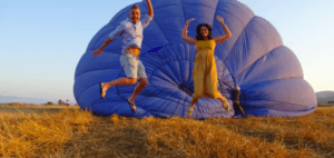 Dos personas saltan alegremente frente a un globo aerostático azul desinflado en un campo de hierba al atardecer, encarnando la esencia mágica de la experiencia Vuelo en globo en Valencia. - ForTwo - Planes para parejas