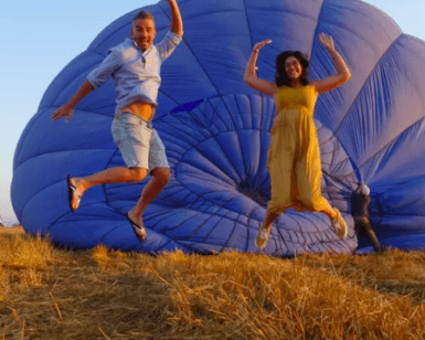 Dos personas saltan alegremente frente a un globo aerostático azul desinflado en un campo de hierba al atardecer, encarnando la esencia mágica de la experiencia Vuelo en globo en Valencia. - ForTwo - Planes para parejas