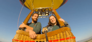 Dos personas sonriendo en una cesta de mimbre de un globo aerostático, disfrutando de un vuelo en globo en Valencia contra el cielo azul claro. - ForTwo - Planes para parejas