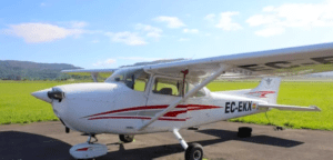 Un pequeño avión blanco con rayas rojas se encuentra en una pista contra campos de hierba y colinas bajo un cielo azul, listo para una aventura con "Vuelo sobre Bilbao y la costa de Bizkaia. - ForTwo - Planes para parejas