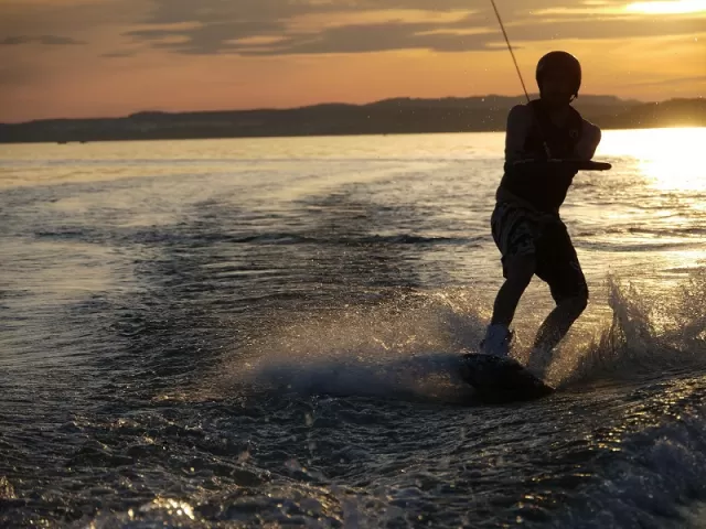 Una persona practica hábilmente el Wakeboard en Gandía sobre las brillantes aguas al atardecer, entre salpicaduras y un vibrante fondo naranja. - ForTwo - Planes para parejas