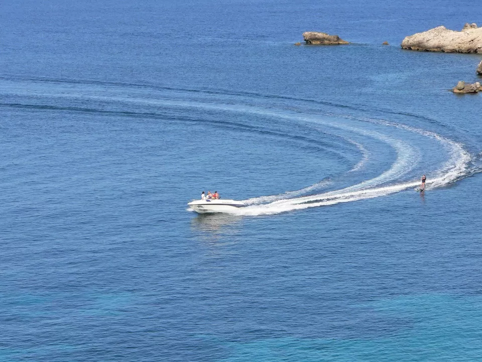 El Wakeboard en Gandía corta el mar azul en calma, arrastrando a un esquiador acuático, mientras crea olas con el telón de fondo de afloramientos rocosos. - ForTwo - Planes para parejas