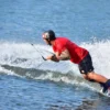 Con camiseta y casco rojos, una persona practica con destreza el "Wakeboard en Gandía" en un pintoresco lago, dejando tras de sí un emocionante rastro de agua pulverizada. - ForTwo - Planes para parejas
