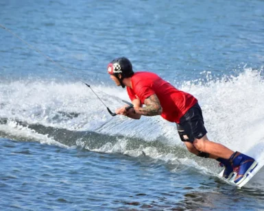 Con camiseta y casco rojos, una persona practica con destreza el "Wakeboard en Gandía" en un pintoresco lago, dejando tras de sí un emocionante rastro de agua pulverizada. - ForTwo - Planes para parejas
