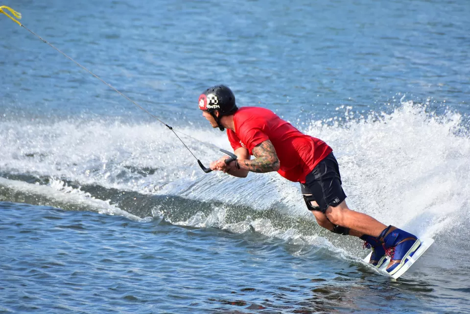Con camiseta y casco rojos, una persona practica con destreza el "Wakeboard en Gandía" en un pintoresco lago, dejando tras de sí un emocionante rastro de agua pulverizada. - ForTwo - Planes para parejas