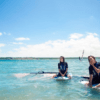 Dos personas se relajan sobre tablas de paddle surf en las tranquilas y cristalinas aguas de la Bahía de Fornells bajo un vibrante cielo azul. Producto: Windsurf en la Bahía de Fornells. - ForTwo - Planes para parejas