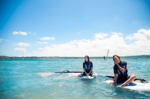 Dos personas se relajan sobre tablas de paddle surf en las tranquilas y cristalinas aguas de la Bahía de Fornells bajo un vibrante cielo azul. Producto: Windsurf en la Bahía de Fornells. - ForTwo - Planes para parejas