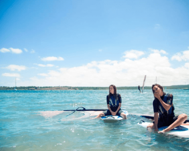 Dos personas se relajan sobre tablas de paddle surf en las tranquilas y cristalinas aguas de la Bahía de Fornells bajo un vibrante cielo azul. Producto: Windsurf en la Bahía de Fornells. - ForTwo - Planes para parejas
