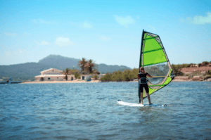 Una persona disfruta del Windsurf en la Bahía de Fornells, deslizándose sobre aguas serenas con montañas lejanas y casas con encanto bajo un cielo azul claro. - ForTwo - Planes para parejas