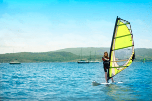 Una persona practicando windsurf con destreza en un lago azul con colinas verdes y barcos en el horizonte, similar a las vistas panorámicas de Windsurf en la Bahía de Fornells. - ForTwo - Planes para parejas