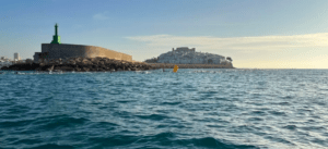 Una escena costera cerca de Alcocéber muestra un rompeolas rocoso y un pueblo distante bajo un cielo despejado, visto a través de aguas azules tranquilas, donde el Yate de lujo en Alcocéber se desliza elegantemente. - ForTwo - Planes para parejas