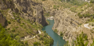 El estrecho río Genil serpentea a través de un cañón rocoso con escasa vegetación y un camino a la derecha, lo que lo convierte en un lugar atractivo para una tranquila experiencia de "kayak por el río Genil" en medio de la agreste belleza de la naturaleza. - ForTwo - Planes para parejas