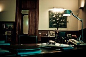 En una oficina con poca luz, que recuerda al Escape Room Misterio del Profesor Waichosky en Zaragoza 🕵️‍♂️, el escritorio de madera de Waichosky está abarrotado de papeles. Una lámpara de tenue luz proyecta sombras sobre un cuadro de Zaragoza, realzando la atmósfera intrigante. - ForTwo - Planes para parejas