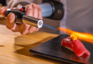 Una persona utiliza expertamente un soplete para dorar sushi en un plato negro, capturando el arte culinario de "Akiro Barcelona: Un Viaje Gastronómico a Japón para Dos". - ForTwo - Planes para parejas
