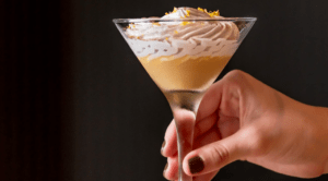 Una mano sostiene un postre en una copa de martini con capas cremosas y ralladura de limón, capturando el espíritu de "Akiro Barcelona: Un Viaje Gastronómico a Japón para Dos" a través de su innovadora fusión de sabores. - ForTwo - Planes para parejas