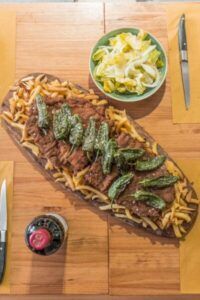 Sobre una rústica mesa de madera se encuentra "El Paraíso de la Carne en Zaragoza", con carne jugosa, papas fritas crujientes y pimientos vibrantes junto a una ensalada fresca y una bebida refrescante: un verdadero festín para el paladar. - ForTwo - Planes para parejas