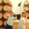 Dos personas celebrando en una bodega con encanto, una de ellas sosteniendo una botella de Cata en Bodega Raíces Ibéricas, con filas de barriles de madera apilados elegantemente en el fondo. - ForTwo - Planes para parejas
