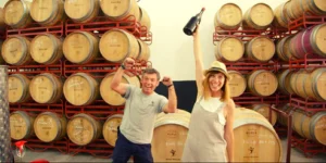 Dos personas celebrando en una bodega con encanto, una de ellas sosteniendo una botella de Cata en Bodega Raíces Ibéricas, con filas de barriles de madera apilados elegantemente en el fondo. - ForTwo - Planes para parejas