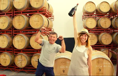 Dos personas celebrando en una bodega con encanto, una de ellas sosteniendo una botella de Cata en Bodega Raíces Ibéricas, con filas de barriles de madera apilados elegantemente en el fondo. - ForTwo - Planes para parejas