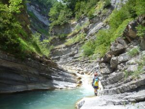 Un excursionista con mochila navega por un cañón rocoso cerca de un arroyo turquesa, enmarcado por una exuberante vegetación, haciendo eco de una ruta aventurera como el "Curso de Vía Ferrata en la Cascada del Sorrosal". - ForTwo - Planes para parejas