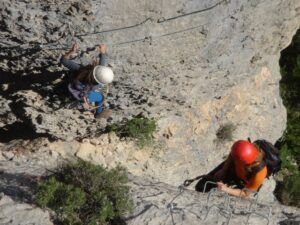 Dos escaladores con casco ascienden por un acantilado rocoso en el emocionante "Curso de Vía Ferrata en la Cascada del Sorrosal", sorteando hábilmente peldaños metálicos y utilizando equipo de seguridad. - ForTwo - Planes para parejas