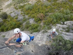 Dos escaladores con casco abordan un acantilado rocoso a lo largo de la vía ferrata en Morata de Jalón, rodeados de exuberante vegetación, con uno más arriba cerca de la cámara. - ForTwo - Planes para parejas