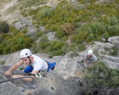 Dos escaladores con casco abordan un acantilado rocoso a lo largo de la vía ferrata en Morata de Jalón, rodeados de exuberante vegetación, con uno más arriba cerca de la cámara. - ForTwo - Planes para parejas