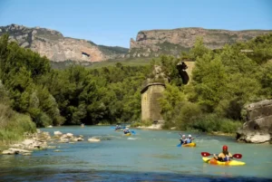 Los kayakistas disfrutan de la emoción del Descenso en Canoa por el Río Gállego, entre árboles verdes y impresionantes montañas rocosas bajo un cielo azul claro. - ForTwo - Planes para parejas
