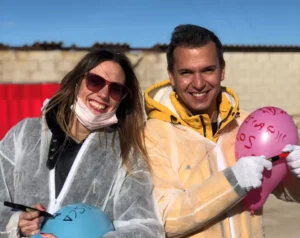 Dos personas con equipo de protección están al aire libre en un soleado día de Zaragoza, sonriendo y sosteniendo globos de colores con texto. Disfrutan de Destructoterapia en Zaragoza, un enfoque terapéutico único que brinda alegría y sanación. - ForTwo - Planes para parejas