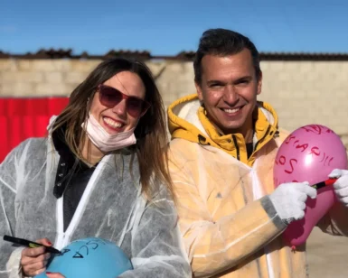 Dos personas con equipo de protección están al aire libre en un soleado día de Zaragoza, sonriendo y sosteniendo globos de colores con texto. Disfrutan de Destructoterapia en Zaragoza, un enfoque terapéutico único que brinda alegría y sanación. - ForTwo - Planes para parejas