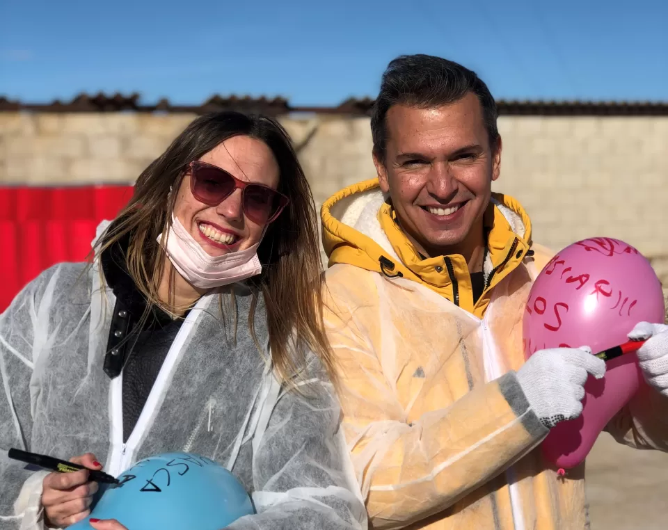 Dos personas con equipo de protección están al aire libre en un soleado día de Zaragoza, sonriendo y sosteniendo globos de colores con texto. Disfrutan de Destructoterapia en Zaragoza, un enfoque terapéutico único que brinda alegría y sanación. - ForTwo - Planes para parejas