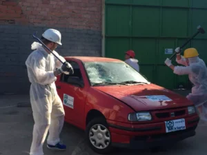 En Zaragoza, tres personas se ponen equipo de protección y liberan estrés destrozando un coche rojo con barras de metal y mazos al aire libre durante la experiencia Destructoterapia en Zaragoza. - ForTwo - Planes para parejas