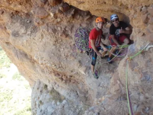Dos escaladores con cascos y cuerdas descansan en la cornisa de un acantilado, saboreando un momento de calma antes de continuar con su Escalada Guiada en Calcena en el accidentado terreno. - ForTwo - Planes para parejas