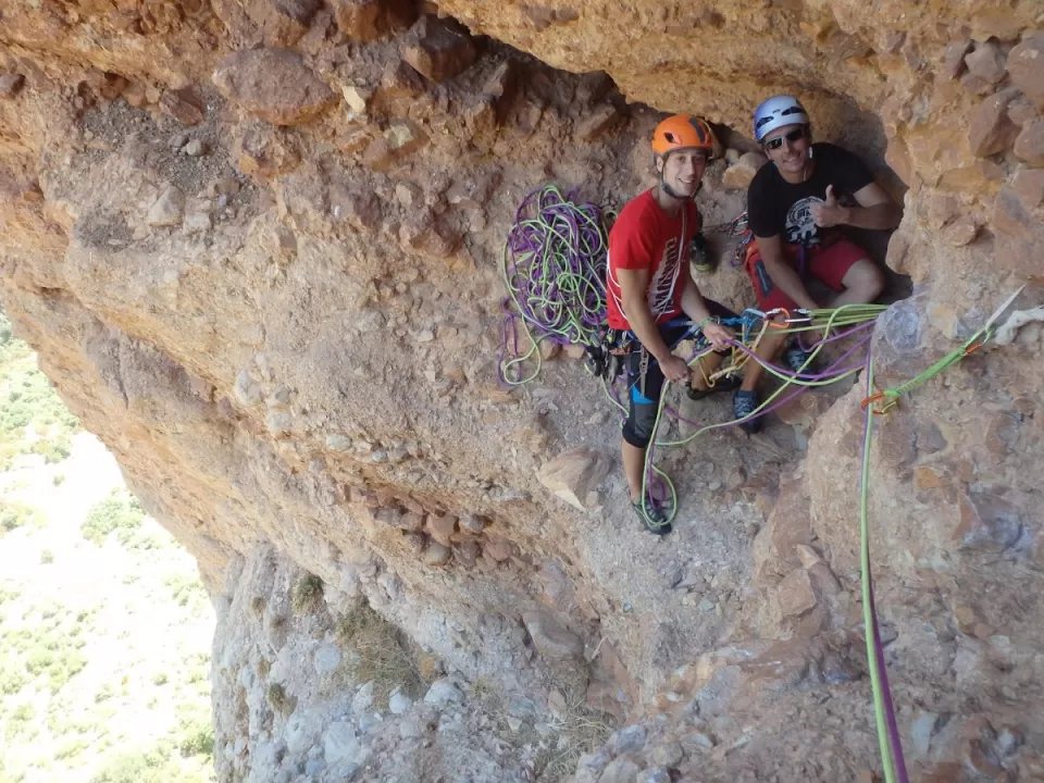 Dos escaladores con cascos y cuerdas descansan en la cornisa de un acantilado, saboreando un momento de calma antes de continuar con su Escalada Guiada en Calcena en el accidentado terreno. - ForTwo - Planes para parejas