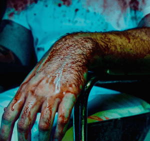 En medio del caos del turno de noche, un brazo ensangrentado reposa sobre una silla metálica. Cerca, una figura con camisa blanca se yergue sobre un entorno manchado, evocando la intensa experiencia de Escape Room Nightshift en Sabadell. - ForTwo - Planes para parejas